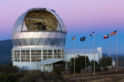 Das Hobby-Eberly Telescope in der Dämmerung. Die Fahnen der vier Partnerländer sind mit der US-Fahne neben der offenen Kuppel zu sehen.