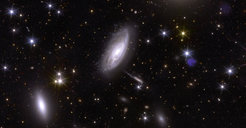 Ein aufgeräumt wirkendes Bild mit elliptisch bläulich leuchtenden Flecken in verschiedenen Größen im Vordergrund, teilweise mit erkennbaren Strukturen, die an einen Strudel erinnern, vor schwarzem Hintergrund mit verteilten scharfen, bläulichen Punkten und gelblichen diffusen Flecken.
