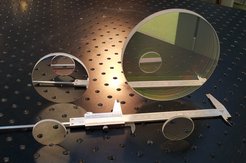 Two small circular discs of the same size stand upright next to each other on a perforated plate, directly behind them in a second row are a disc about twice as large and a disc four times as large. A calliper lies between the two rows. The discs reflect what is in front of them.
